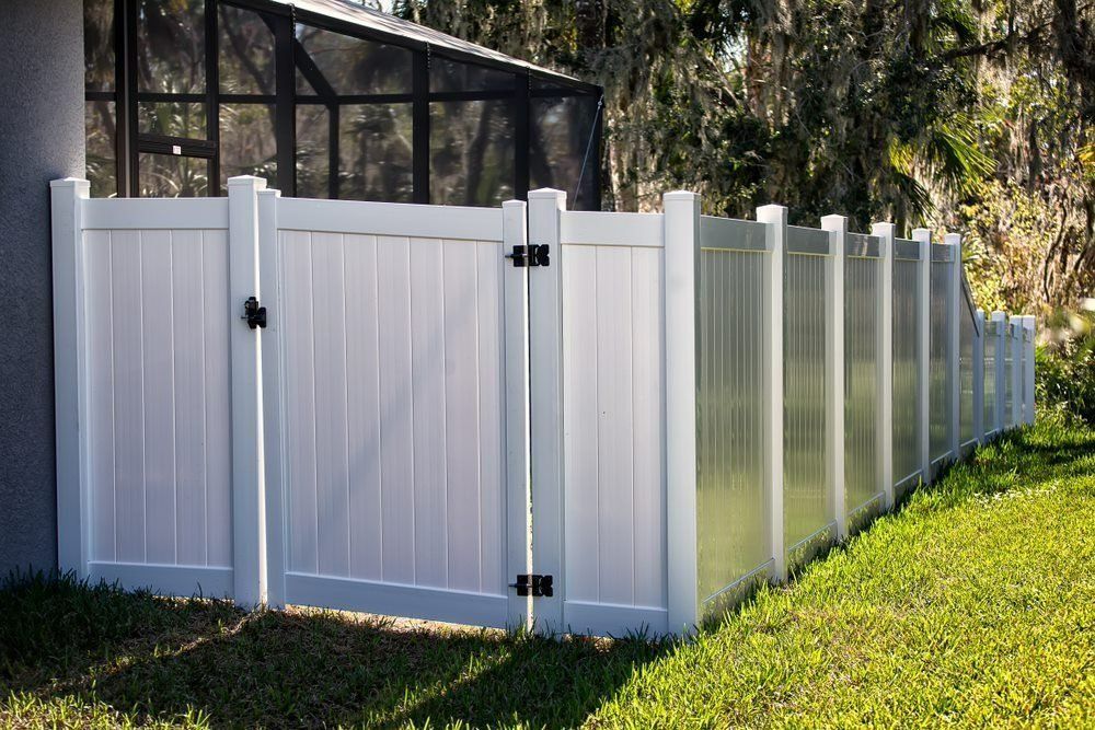 A White Fence With A Black Gate Is Sitting In The Grass Next To A House — Rabbo's Fencing Services In Pinelands, NT