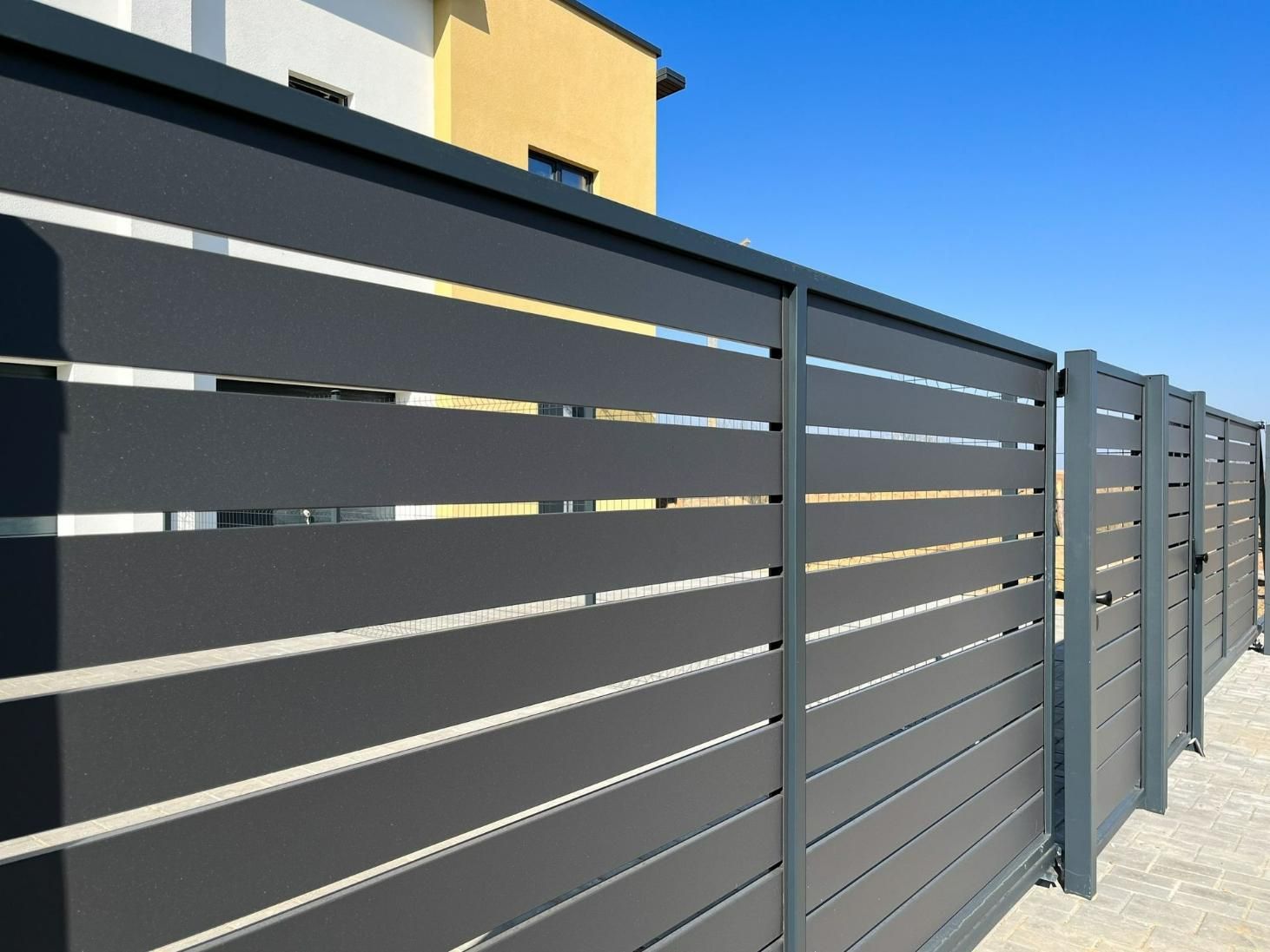 A Gray Fence With A Yellow Building In The Background — Rabbo's Fencing Services In Pinelands, NT