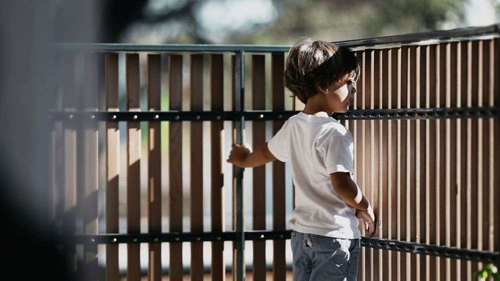 A Young Boy Is Standing Next To A Wooden Fence — Rabbo's Fencing Services In Pinelands, NT