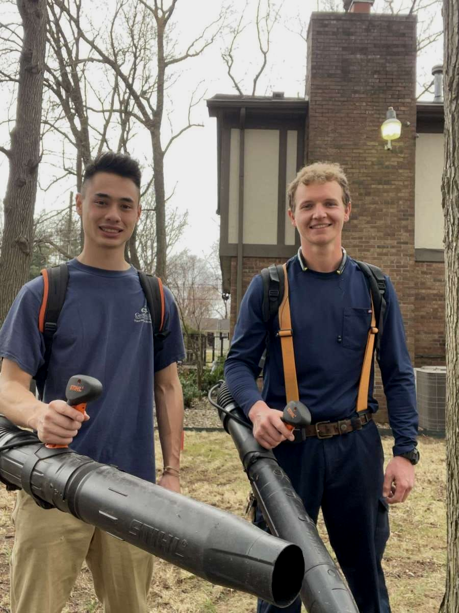 Two people in work clothes holding leaf blowers in front of a brick building.