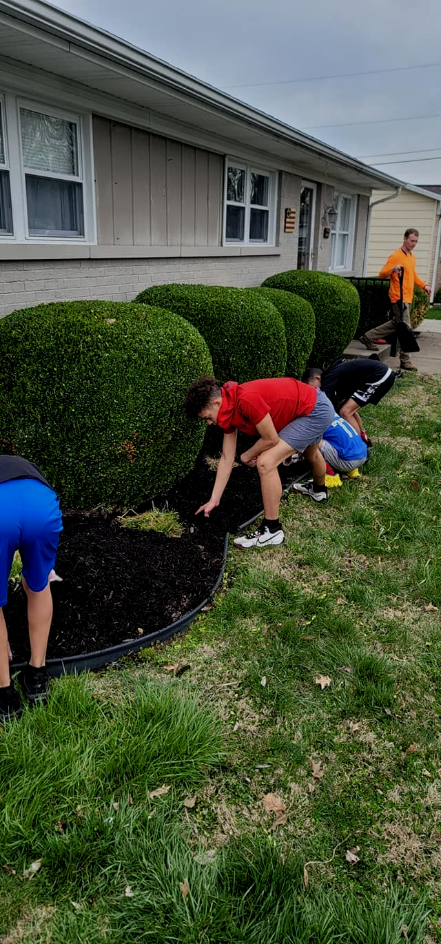 People mulching a flower bed in front of a house.