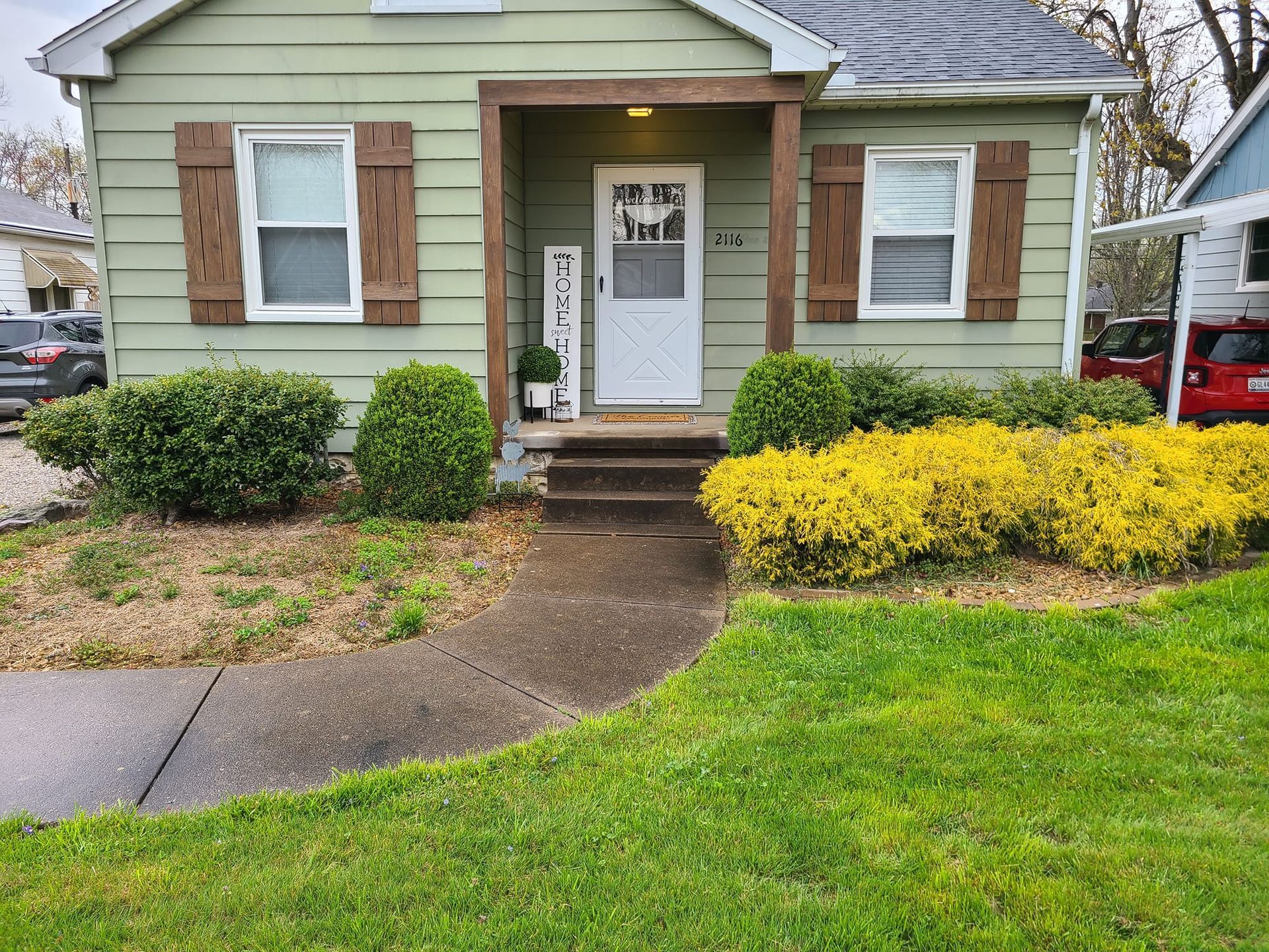 Small green house with brown shutters and a walkway to the front door, landscaped with shrubs and grass.