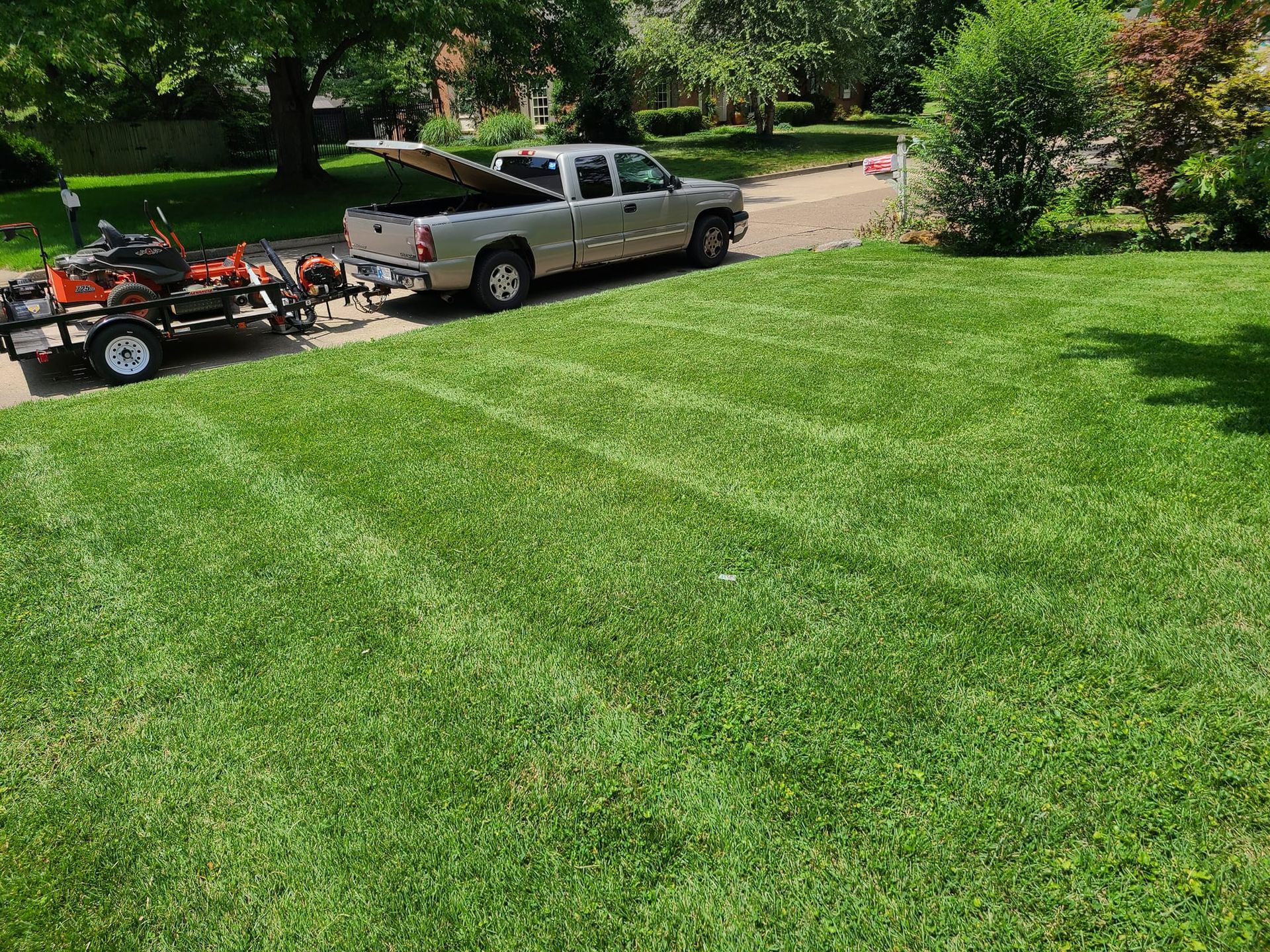 Truck with open bed and trailer carrying lawnmower on freshly mowed lawn, in residential area.