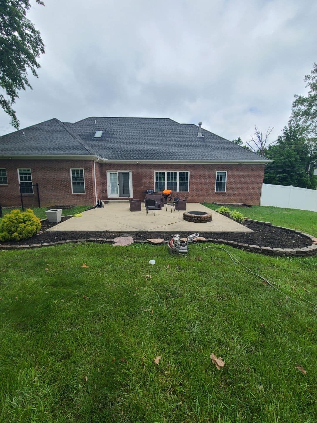 Backyard patio with fire pit, grill, and chairs in front of a brick house with green grass.