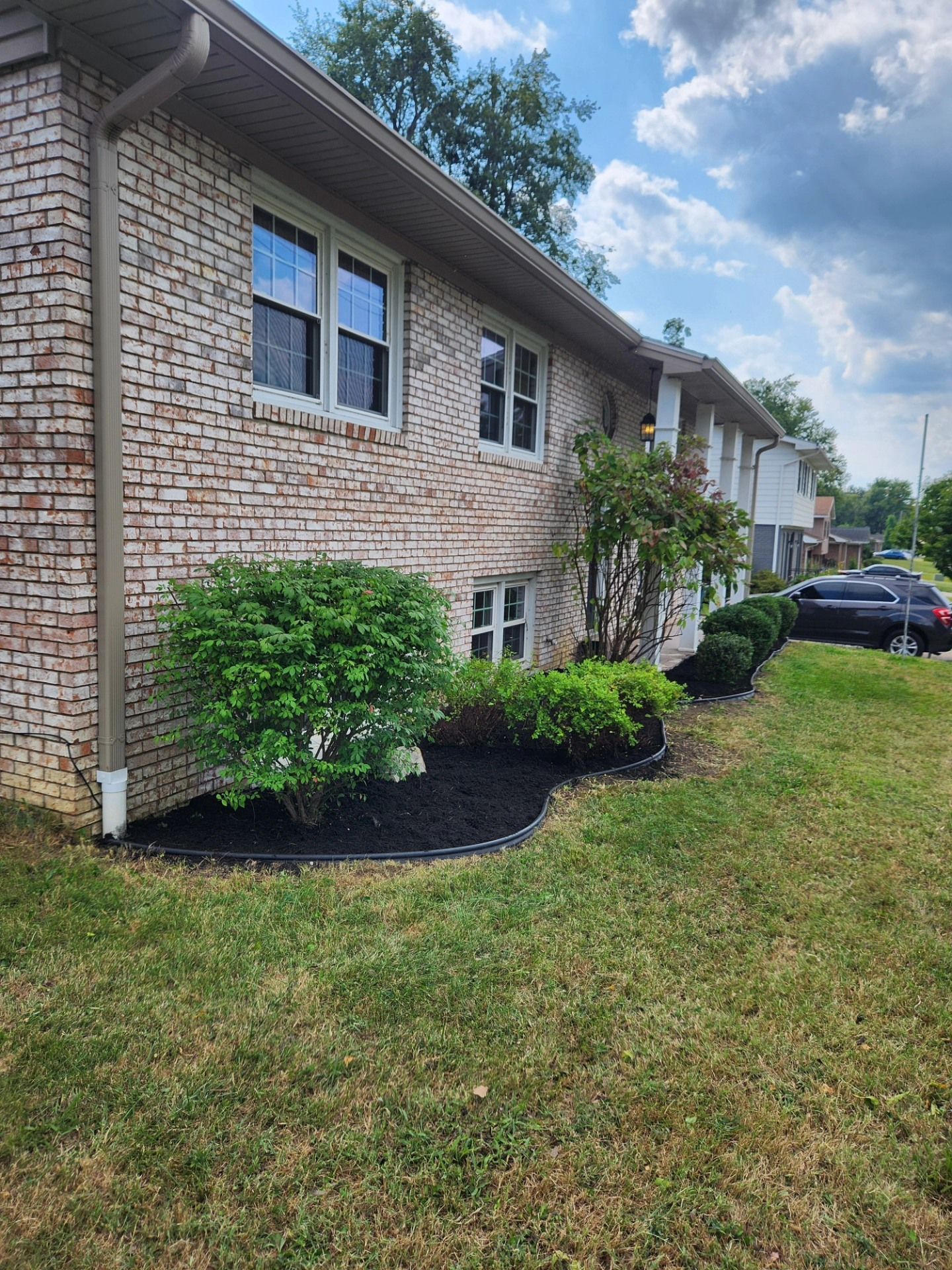 Brick apartment building with black mulch beds, green bushes, and a grassy lawn under a partly cloudy sky.