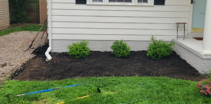 A house with a newly mulched flower bed and three green bushes. Grass and a gravel path are also visible.