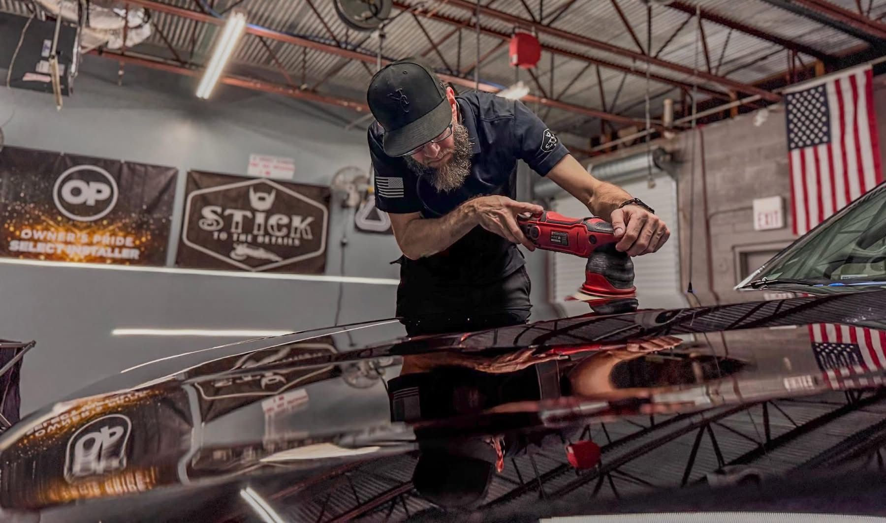 Detailing a black car hood with a red polisher in an auto shop, banners and an American flag in background