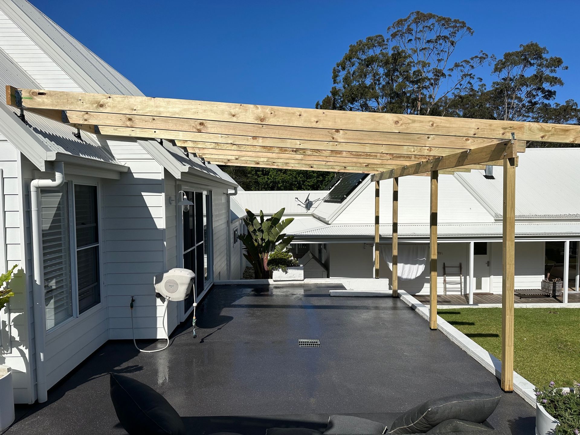 A wooden pergola attached to a white house with a dark, textured deck overlooking a green backyard on a sunny day.