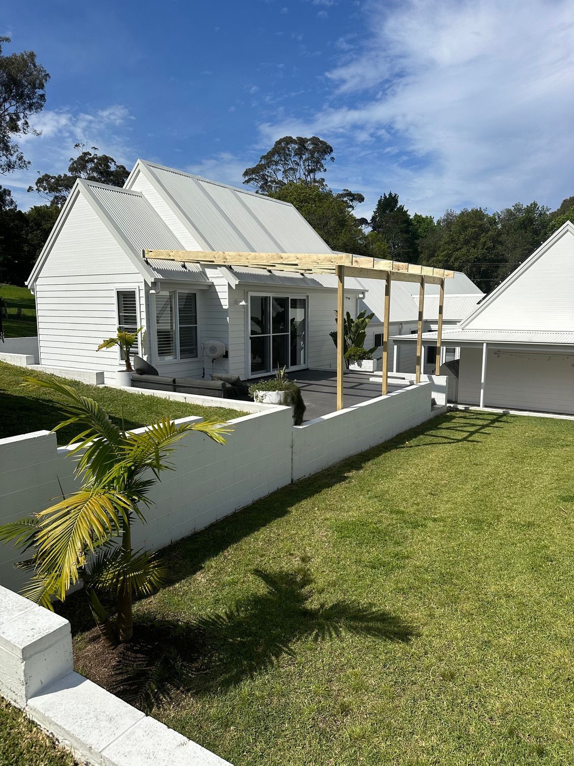 A white house with a metal roof and a wooden pergola frame, set in a sunny backyard with a green lawn and white wall.