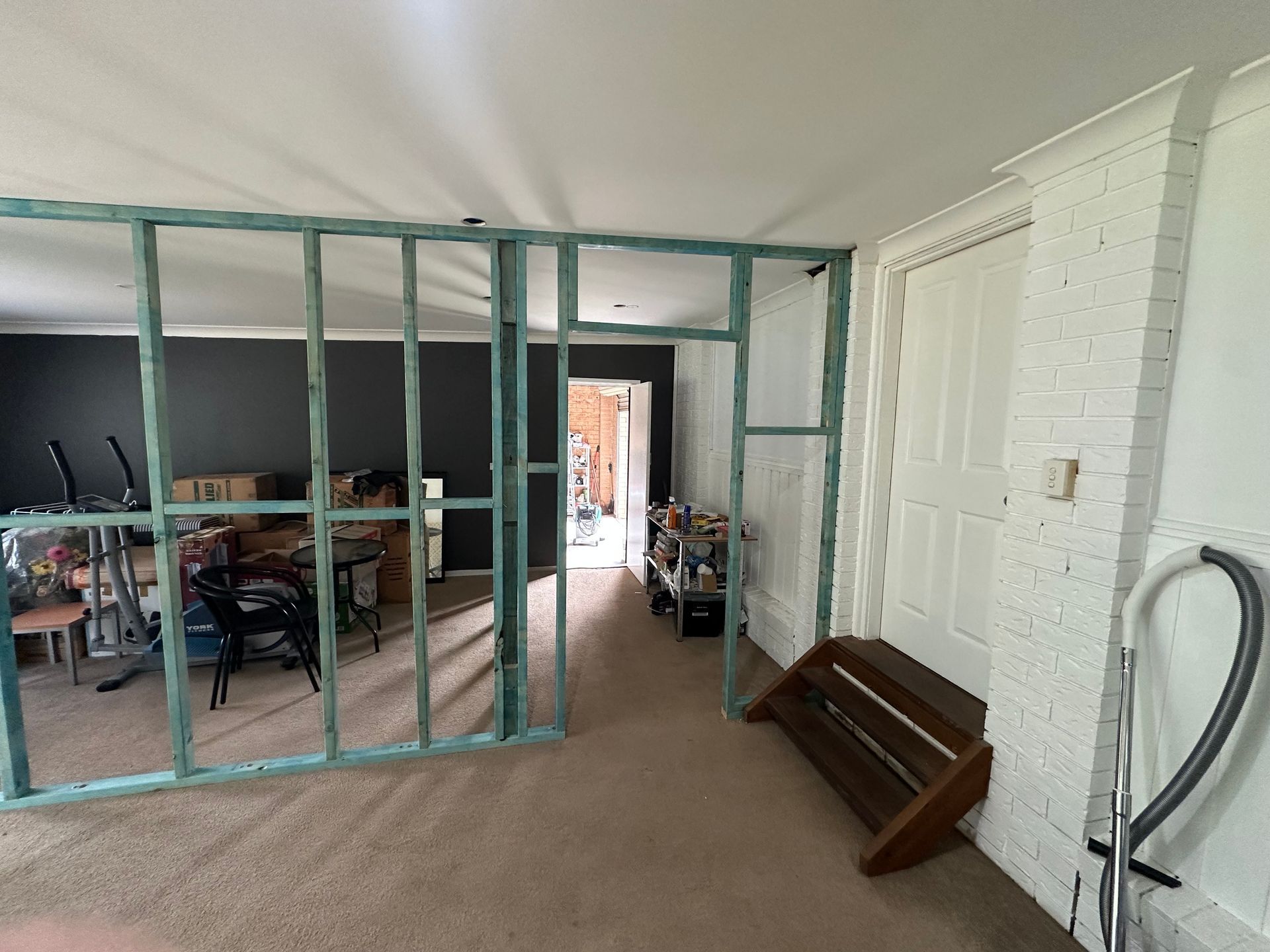 Framed wall studs create a room partition in a space with beige carpet, a white door, and a white brick fireplace pillar.