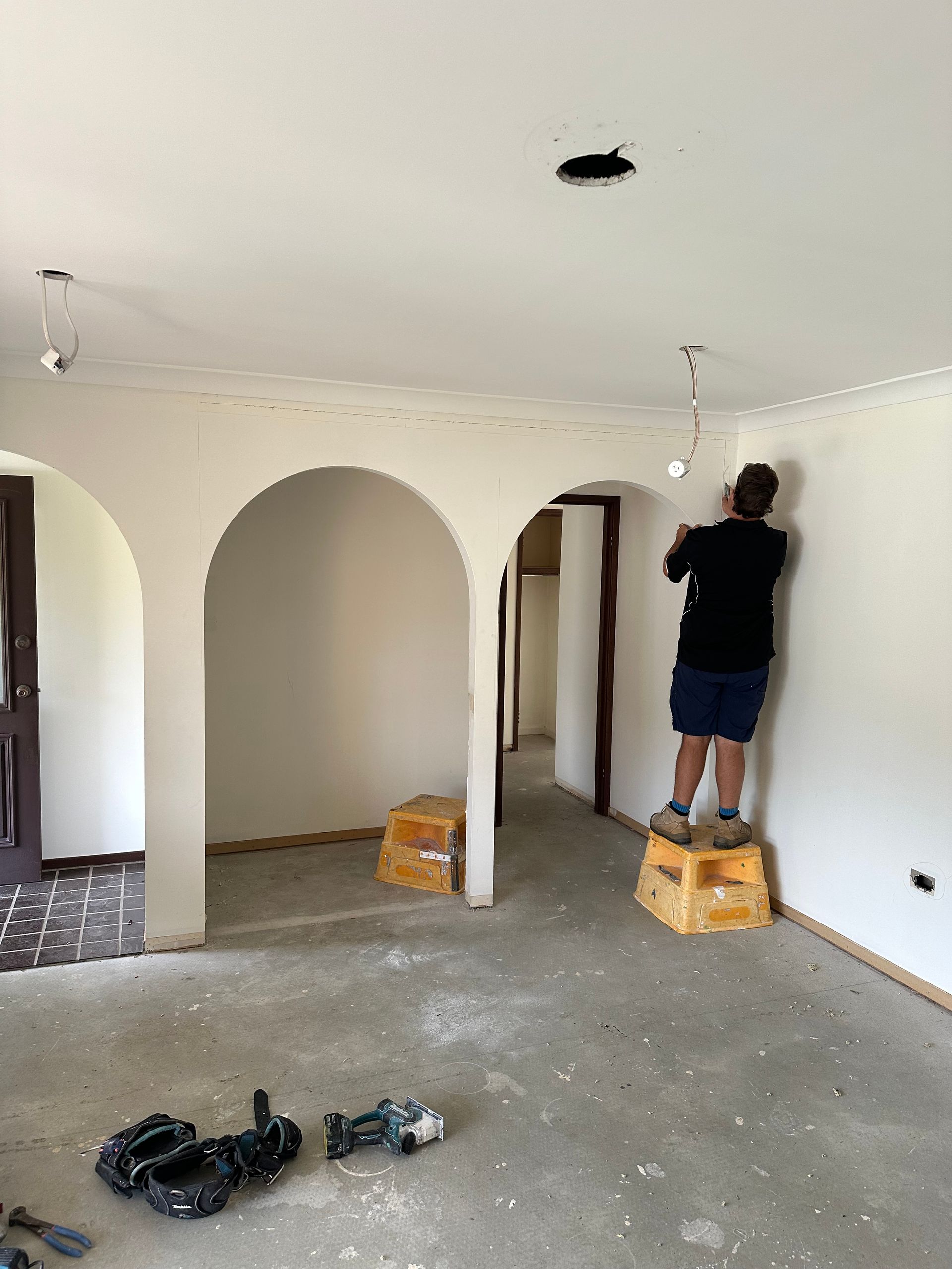 Person standing on a box painting a white room with arched doorways and scattered tools on the floor