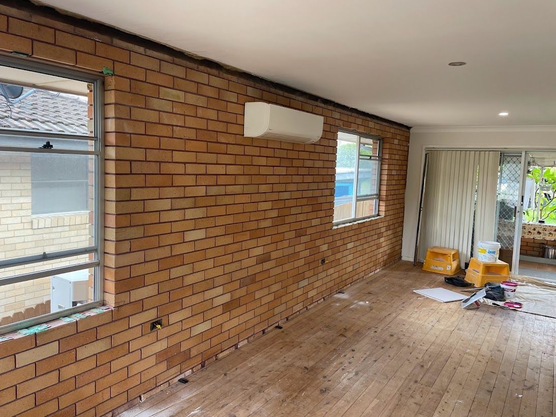 A room under renovation featuring an exposed brick wall, a wall-mounted air conditioner, and wooden flooring.