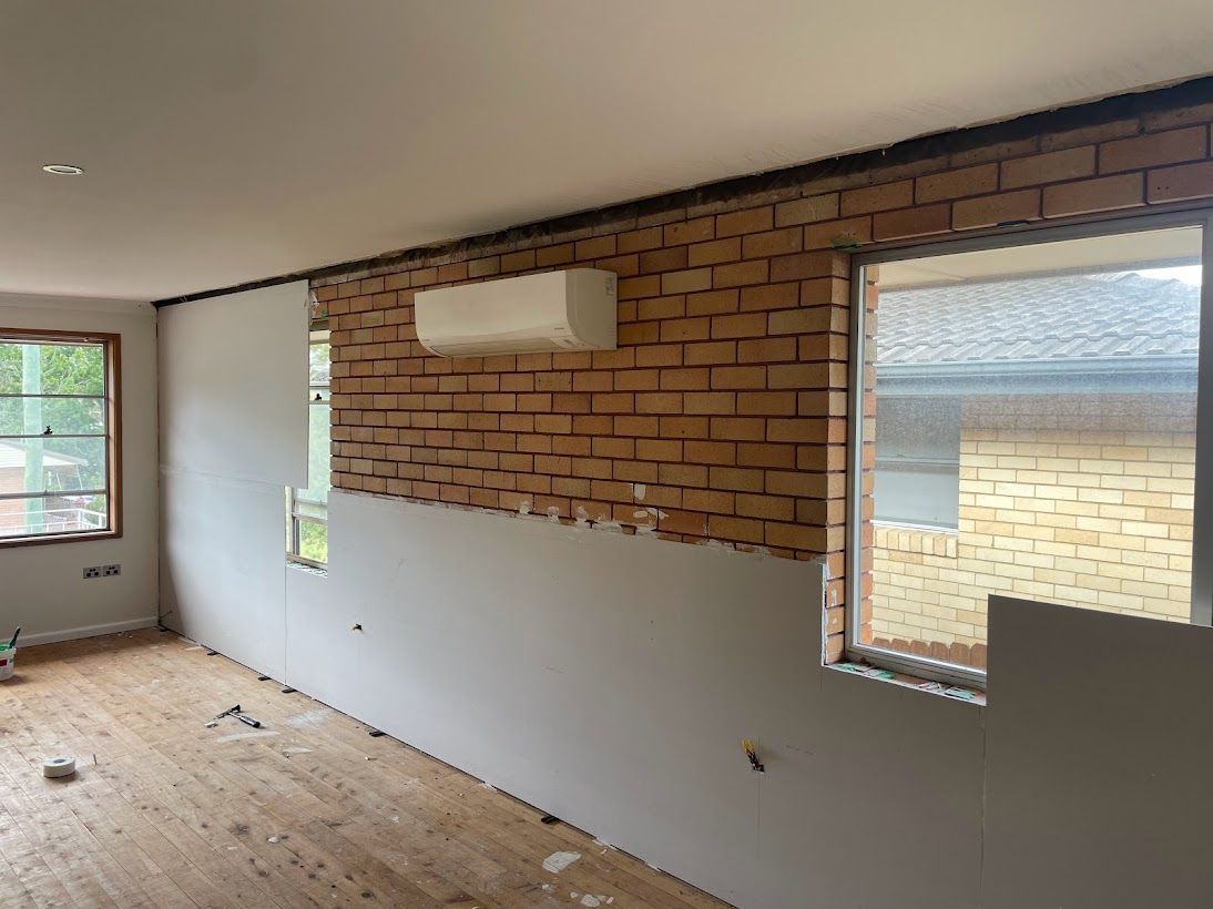 Interior view of a room under renovation with a partially exposed brick wall, drywall panels, and a mounted air conditioner.