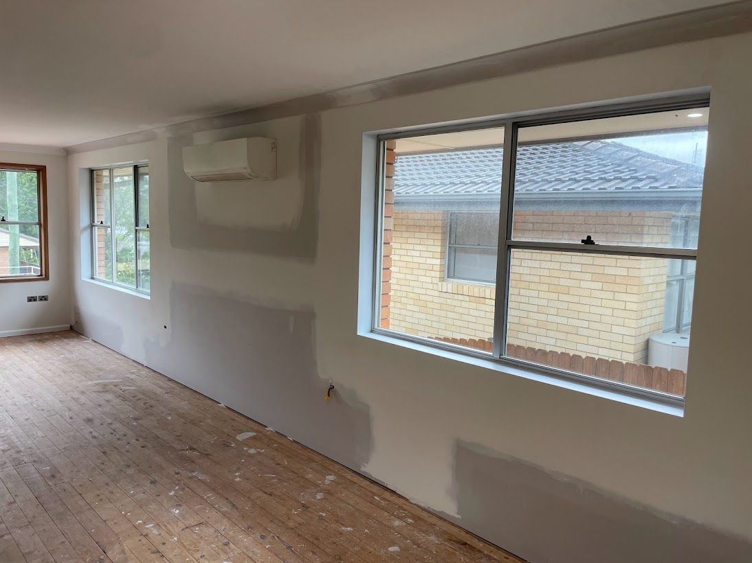 Interior view of a room under renovation with freshly plastered walls, exposed wooden floors, and windows.