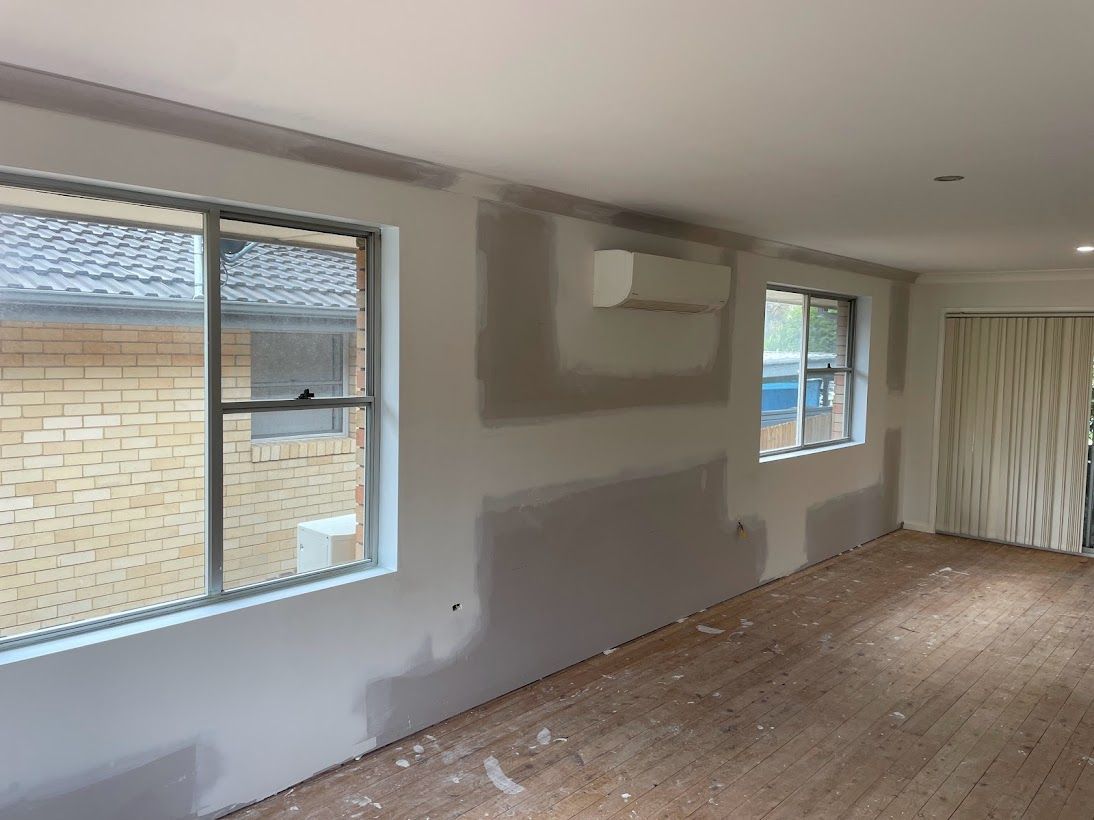 A room undergoing renovations with fresh drywall patches on the walls, two windows, and an air conditioning unit.