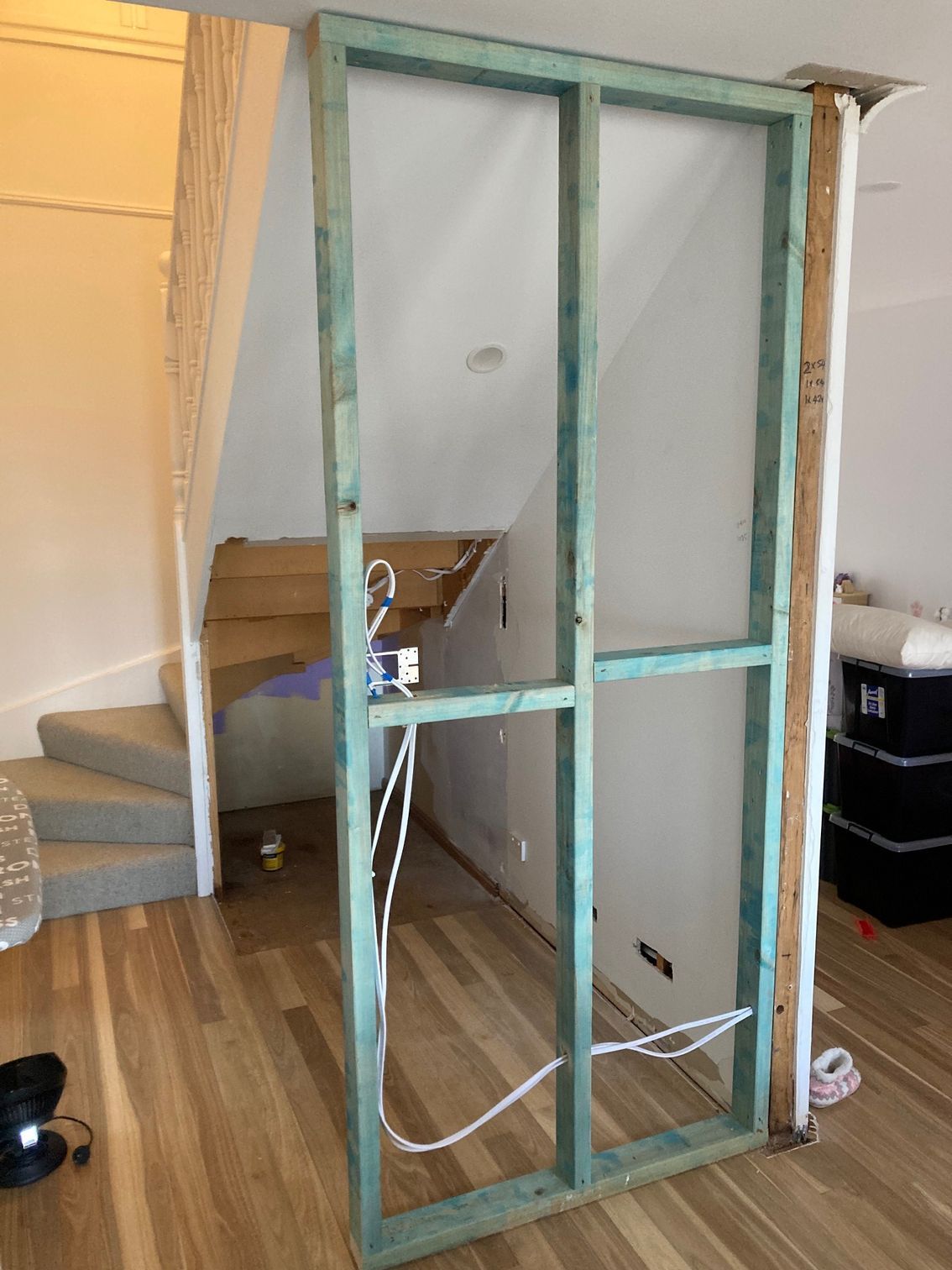A green wooden wall frame stands in a home with wooden floors, with stairs in the background and exposed electrical wiring.