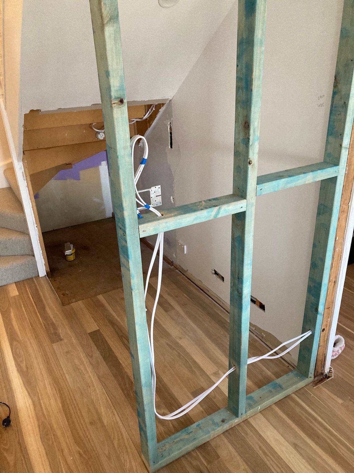 A wooden wall frame under construction stands on a hardwood floor near an open staircase with exposed wiring.