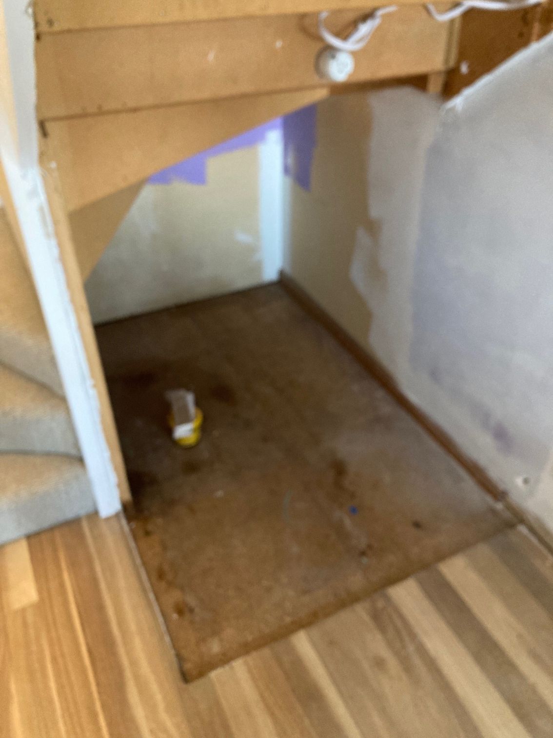 An unfinished alcove under a staircase with exposed wood floorboards, partially painted walls, and a small paint container.