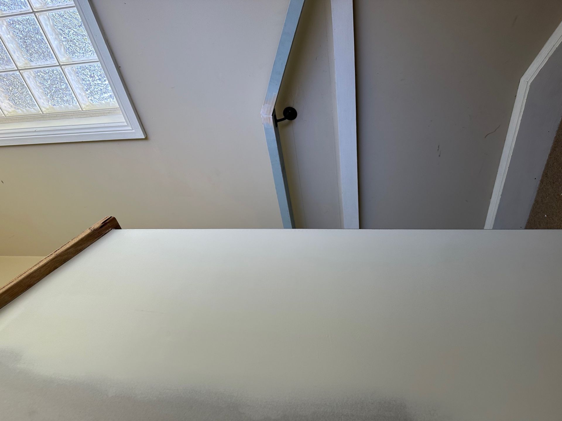 A high-angle view of a white, angled interior wall next to a wooden trim piece, near a window with glass blocks.