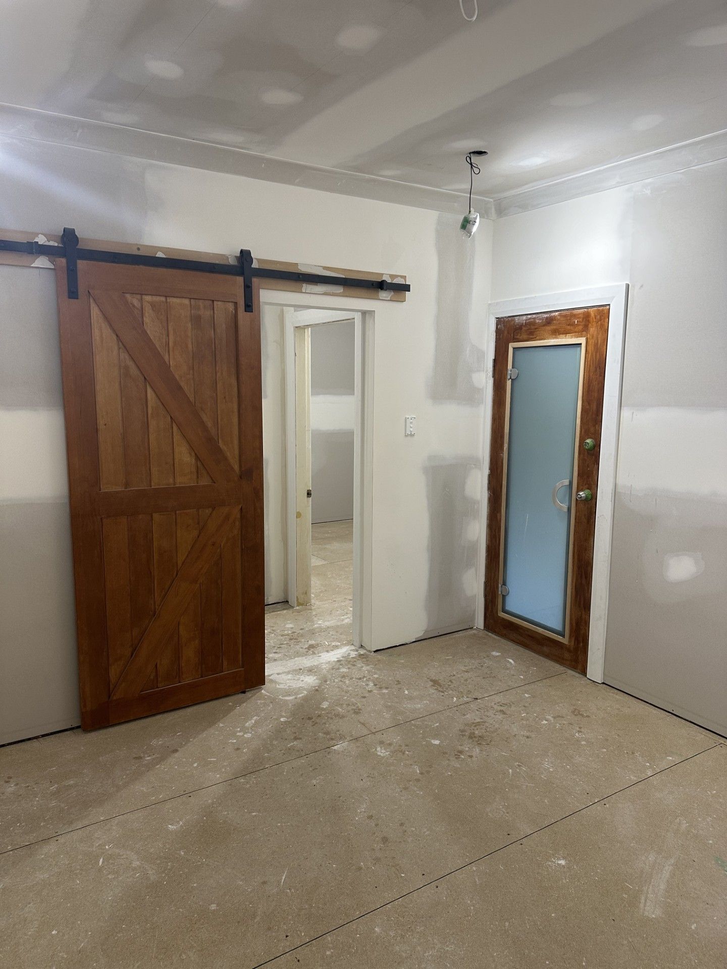 A room under construction with a wooden barn door, an open doorway leading to another area, and a glass-paneled door.