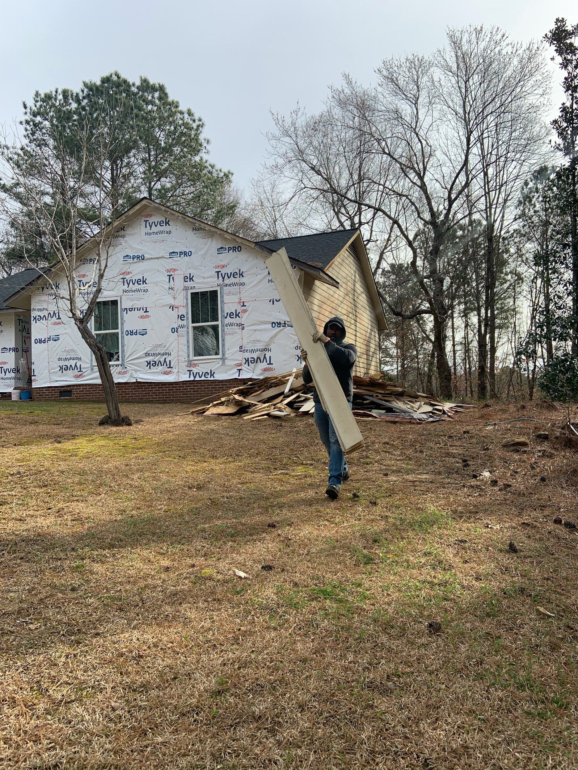 Person carrying a long board near a house under construction, surrounded by trees.