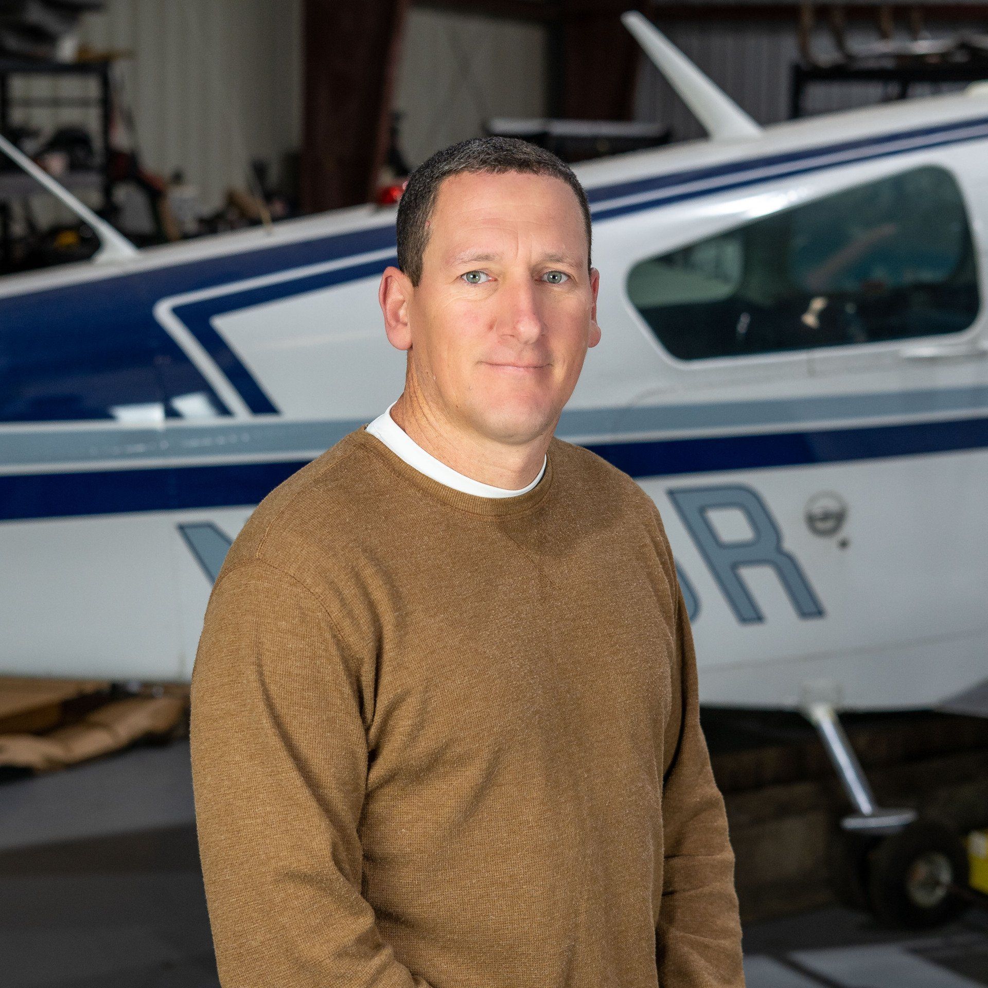 A man in a brown sweater stands in front of a plane with the letter r on it