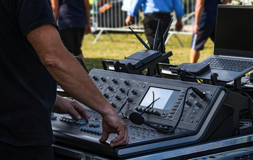 Person operating a sound mixing console outdoors, adjusting knobs and sliders.