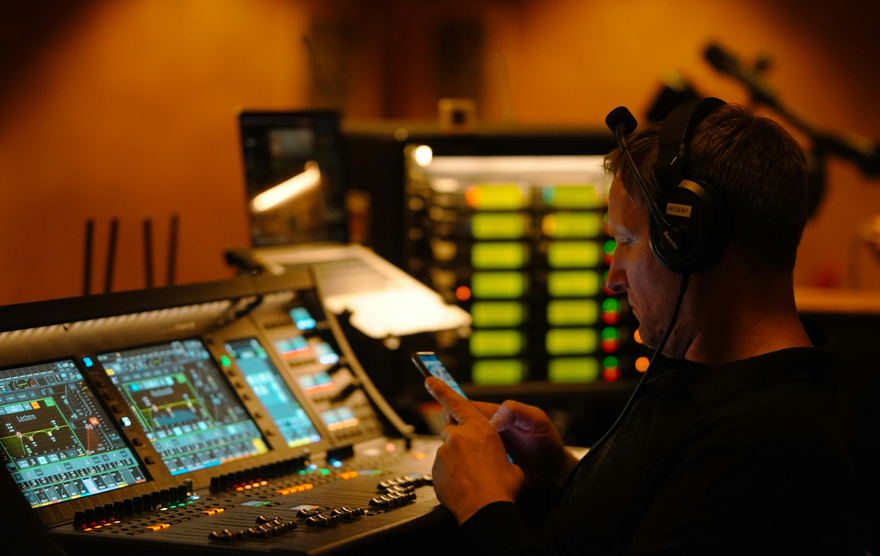Man wearing headphones uses a phone while sitting at a sound mixing console with multiple screens and buttons.