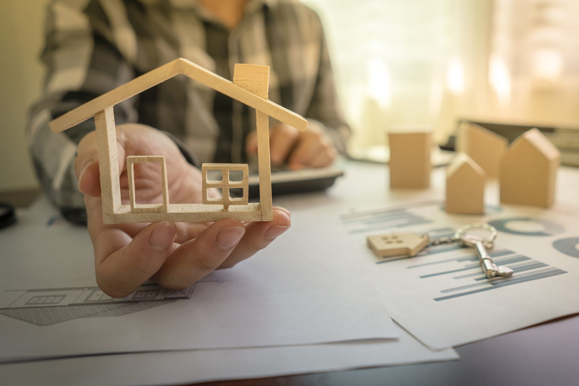 Hands holding a small wooden house model over property plans and keys on a desk