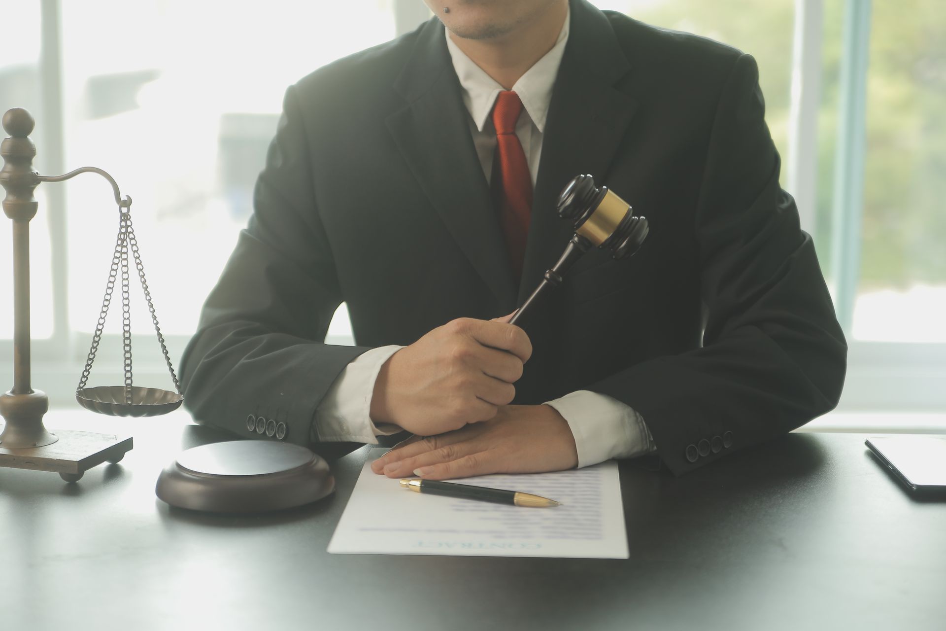 Person in suit holding gavel, scales of justice and document on desk.