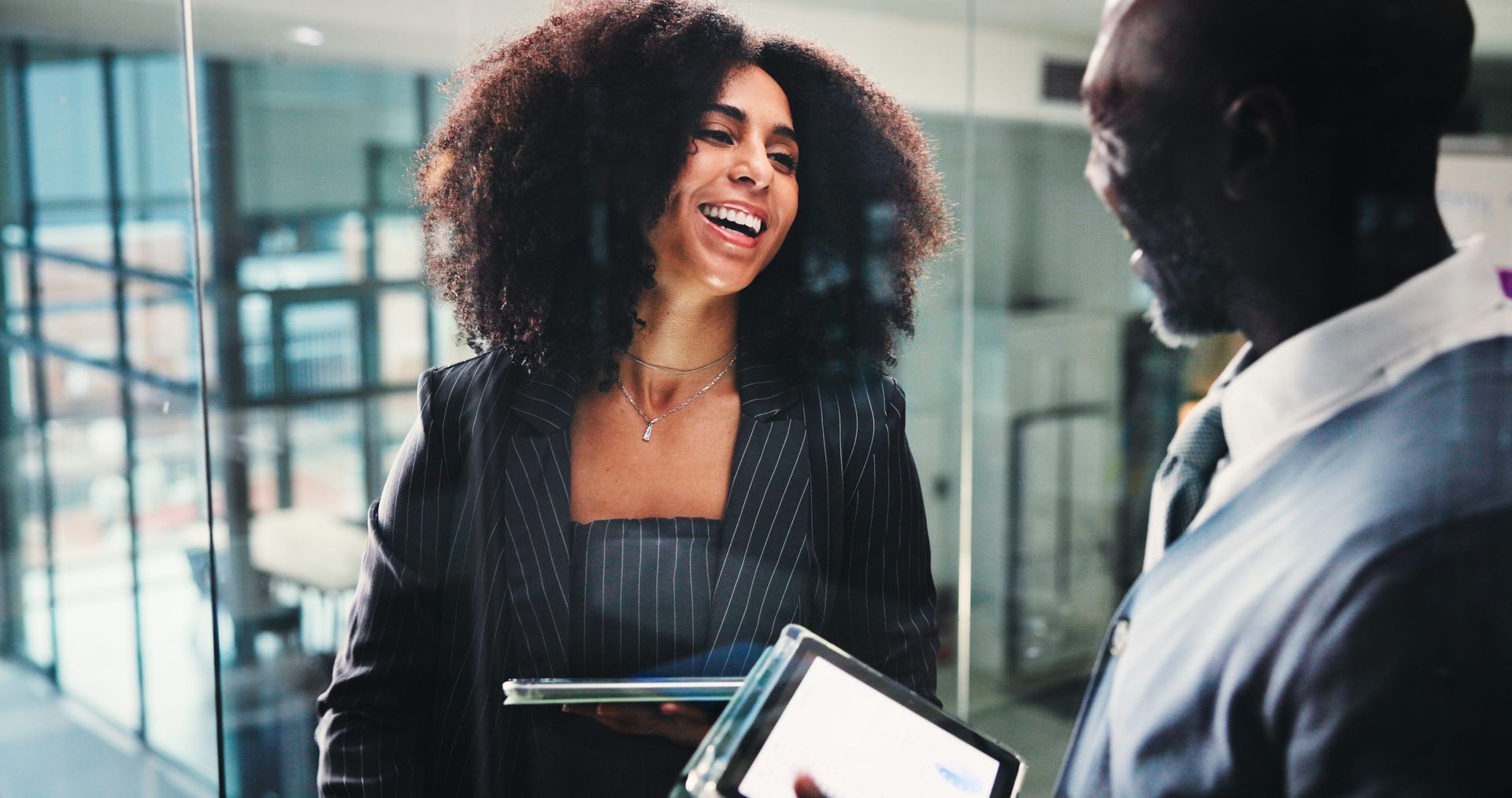 Two people smiling during a business meeting in a modern office, one holding a tablet.