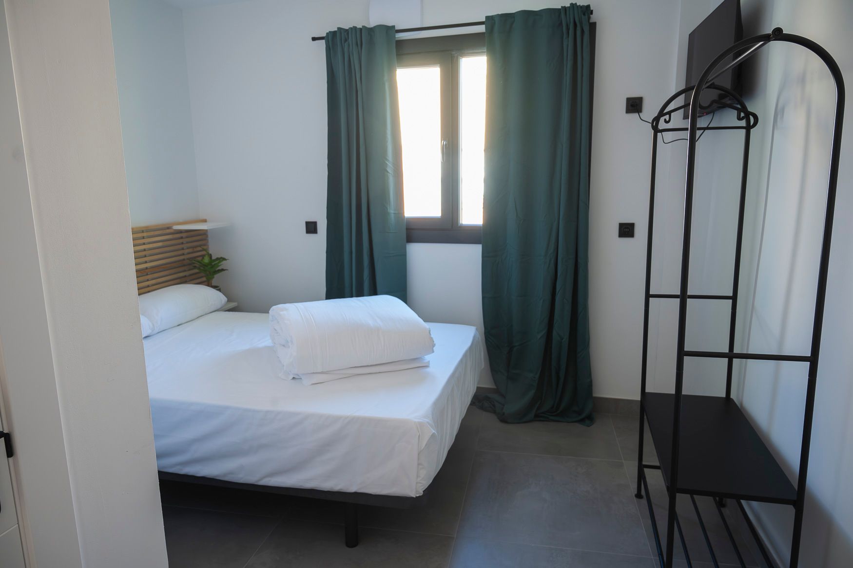 Pequeño dormitorio con una cama, ropa de cama blanca y una ventana con cortinas beige.
