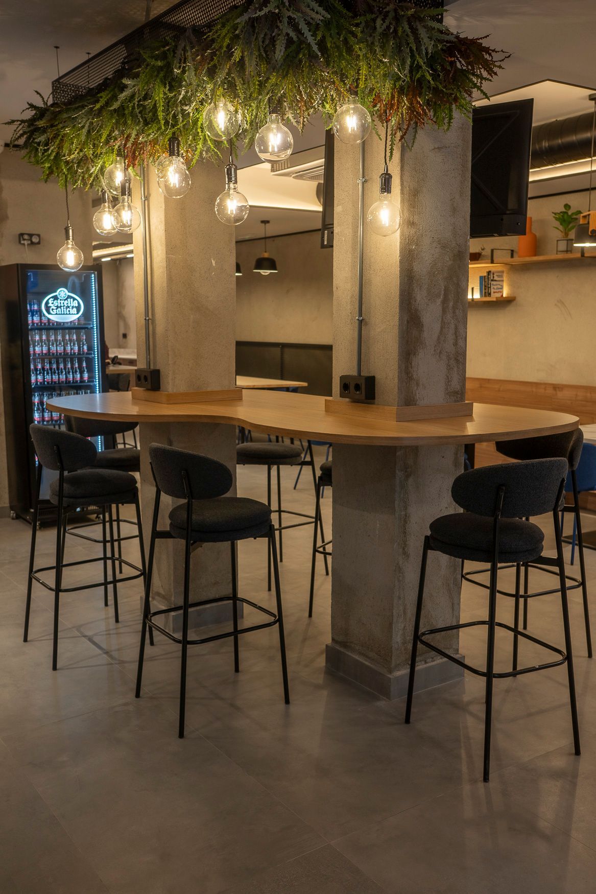 Interior de restaurante con mesas altas, taburetes de bar y luces colgantes sobre un comedor común.