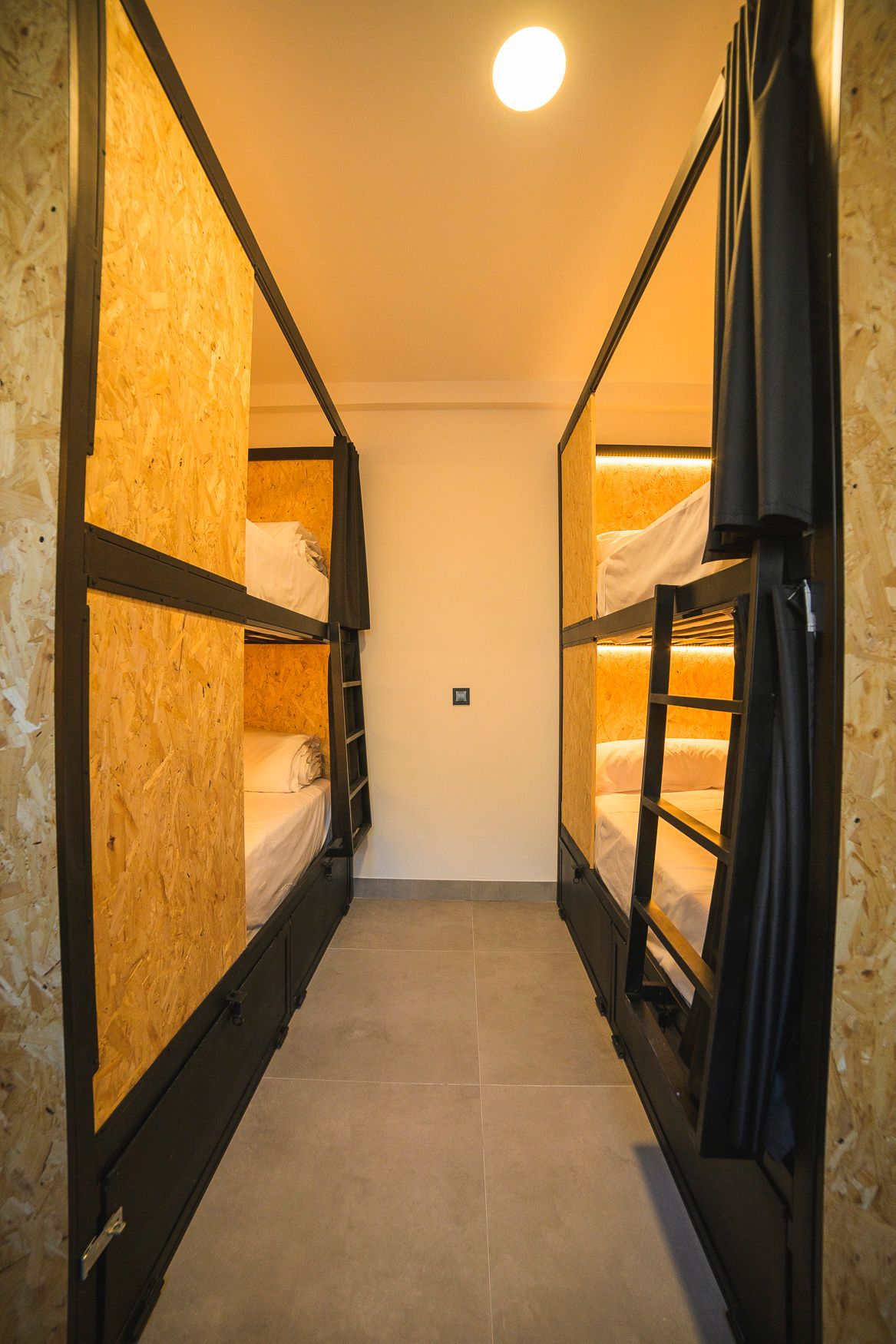 Habitación estrecha con dos literas, cada una con dos camas. Estructura de madera y metal negro.