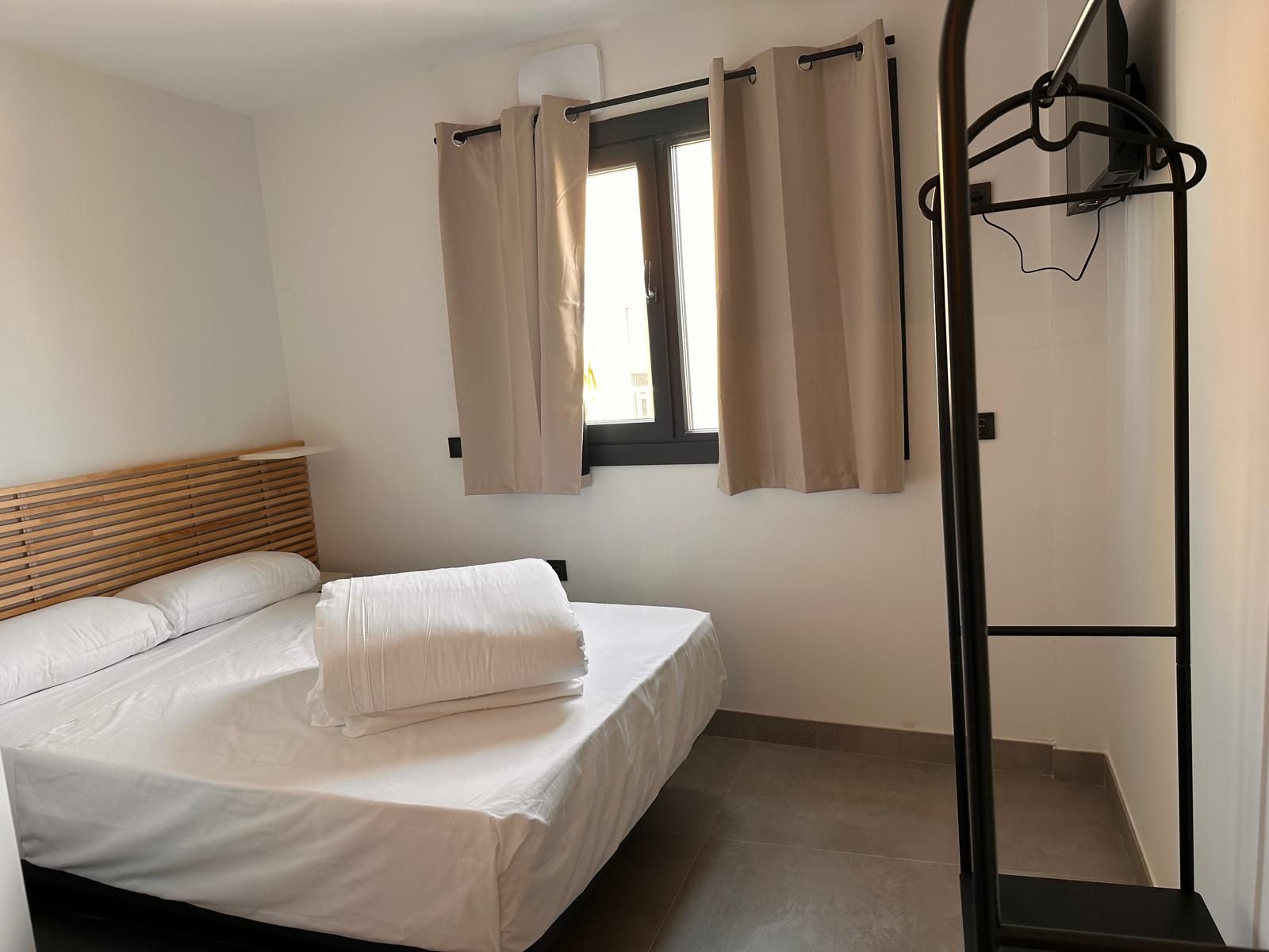 Pequeño dormitorio con cama de matrimonio, paredes claras y ventana con cortinas beige.