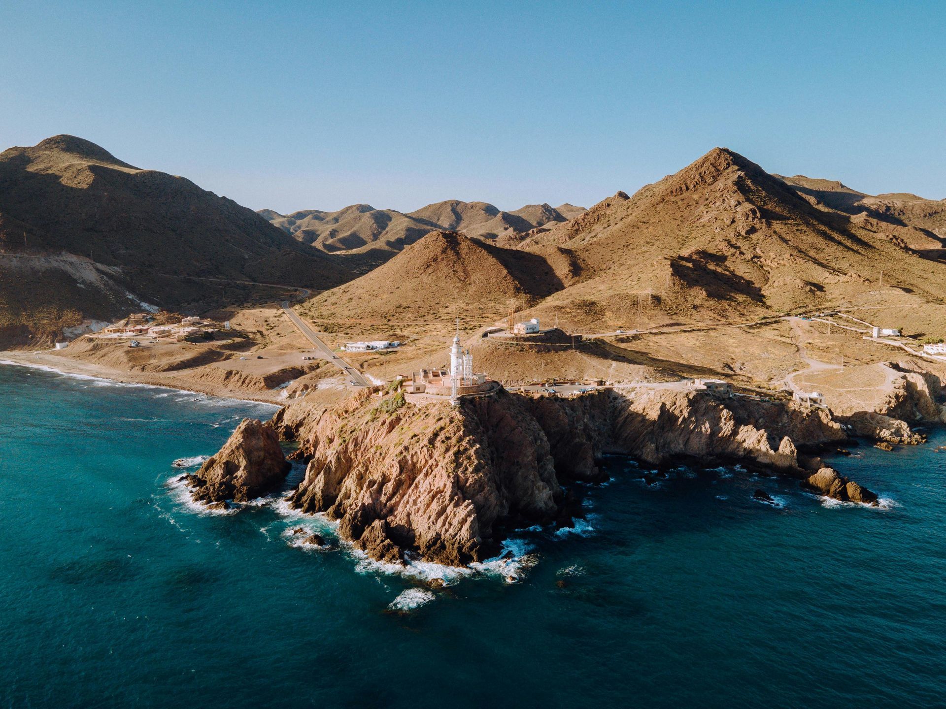 Faro en una punta rocosa, agua turquesa, montañas marrones y cielo despejado.