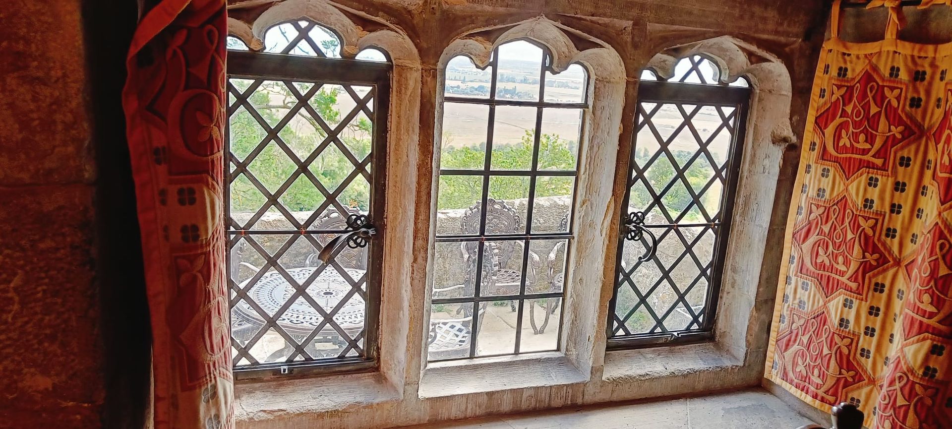 Stone window with three diamond-paned panes, framed by red curtains and patterned fabric walls.