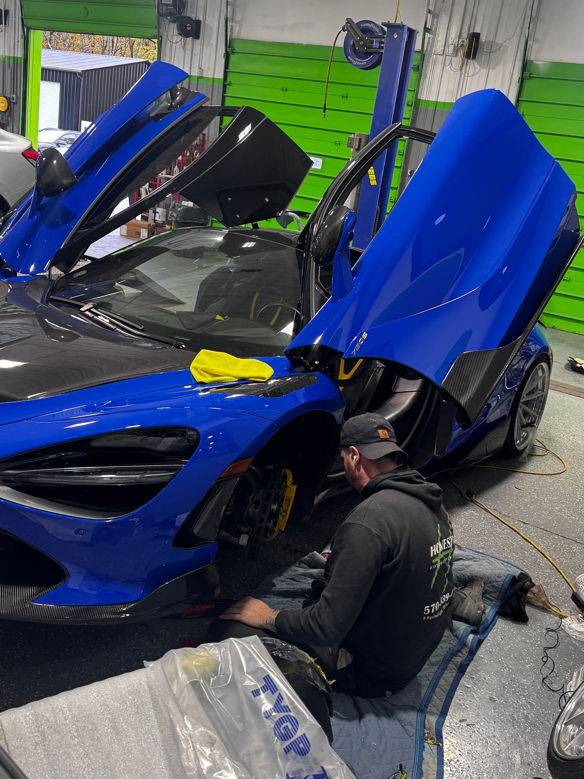 Blue McLaren sports car with doors open, being worked on in a garage by a person. | Honesty Automotive Repairs