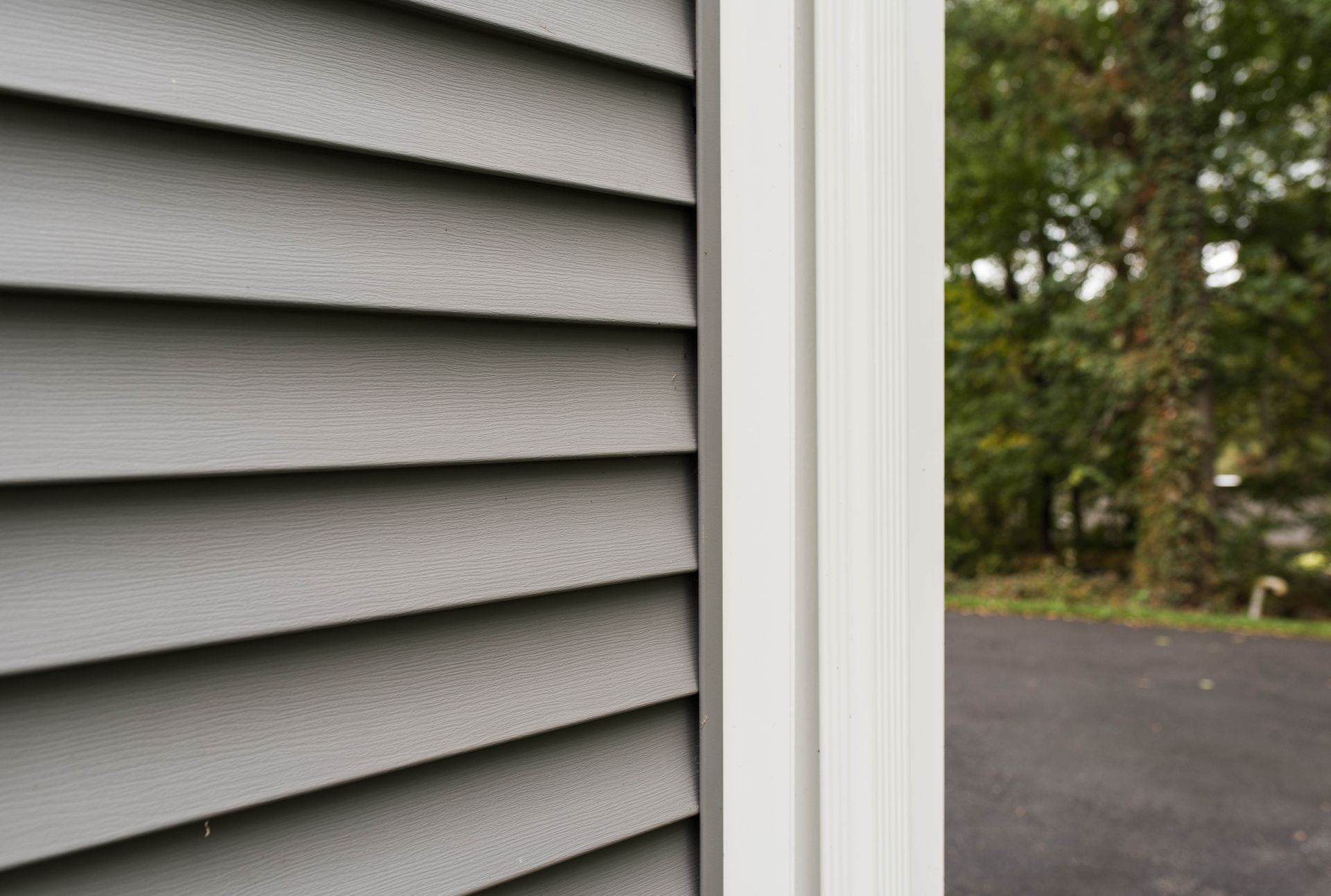 Gray house siding beside a white trim frame, with a driveway and trees in the background
