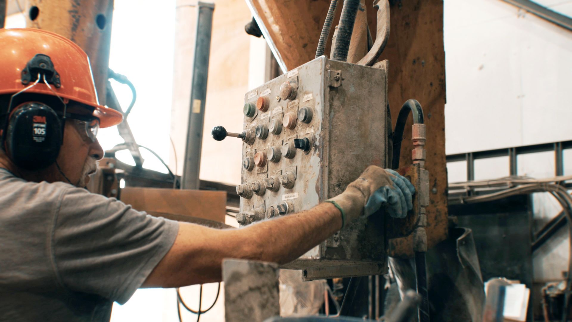 Worker in orange hard hat operating a control panel beside industrial machinery and wooden beams.