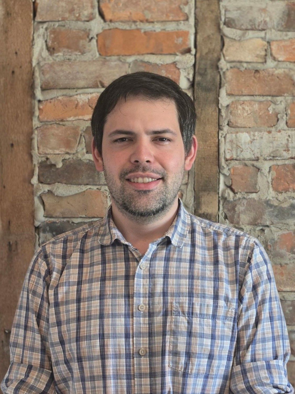 A man in a suit and blue shirt is smiling in front of a brick wall.