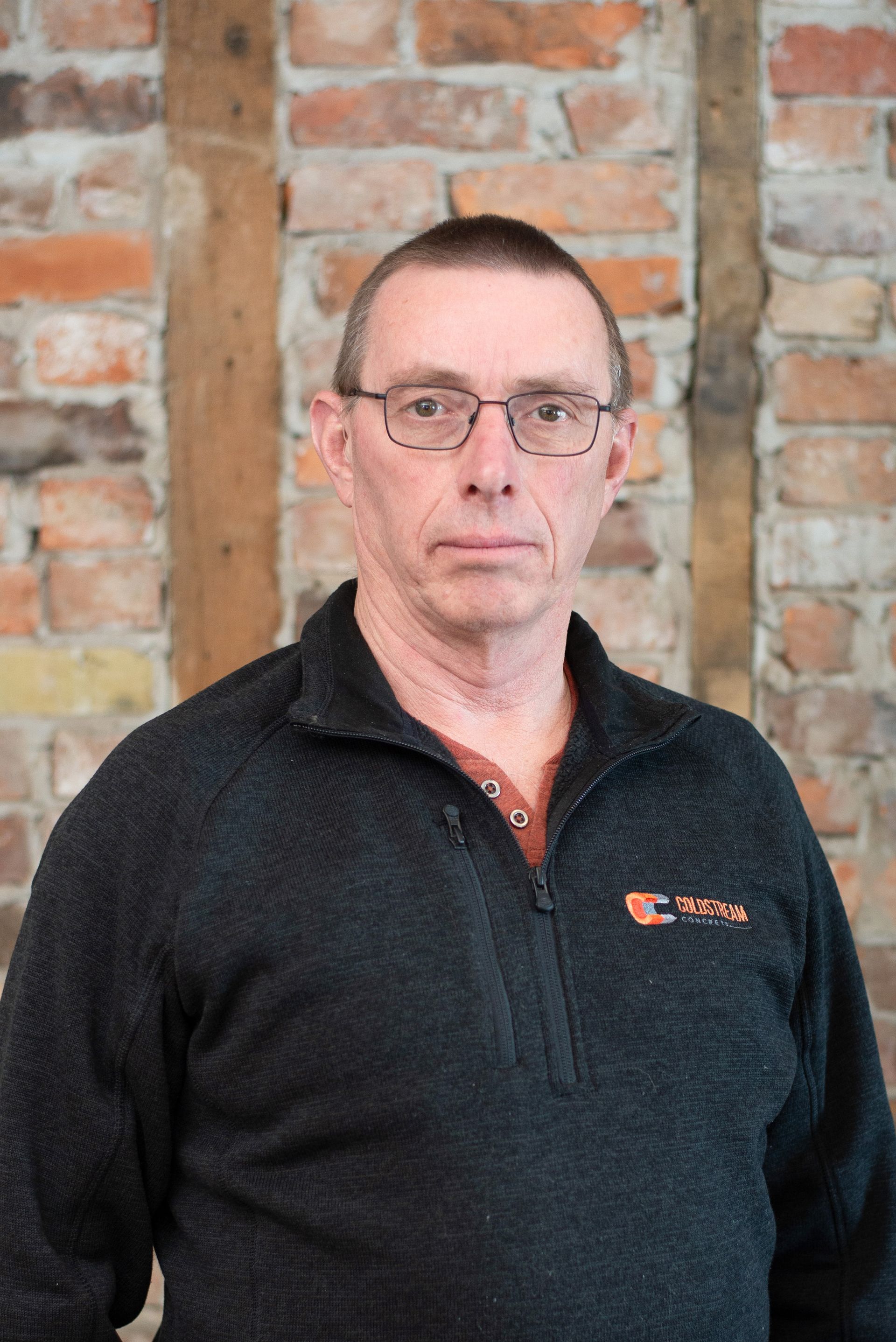 A man with a beard and a black jacket is standing in front of a brick wall.
