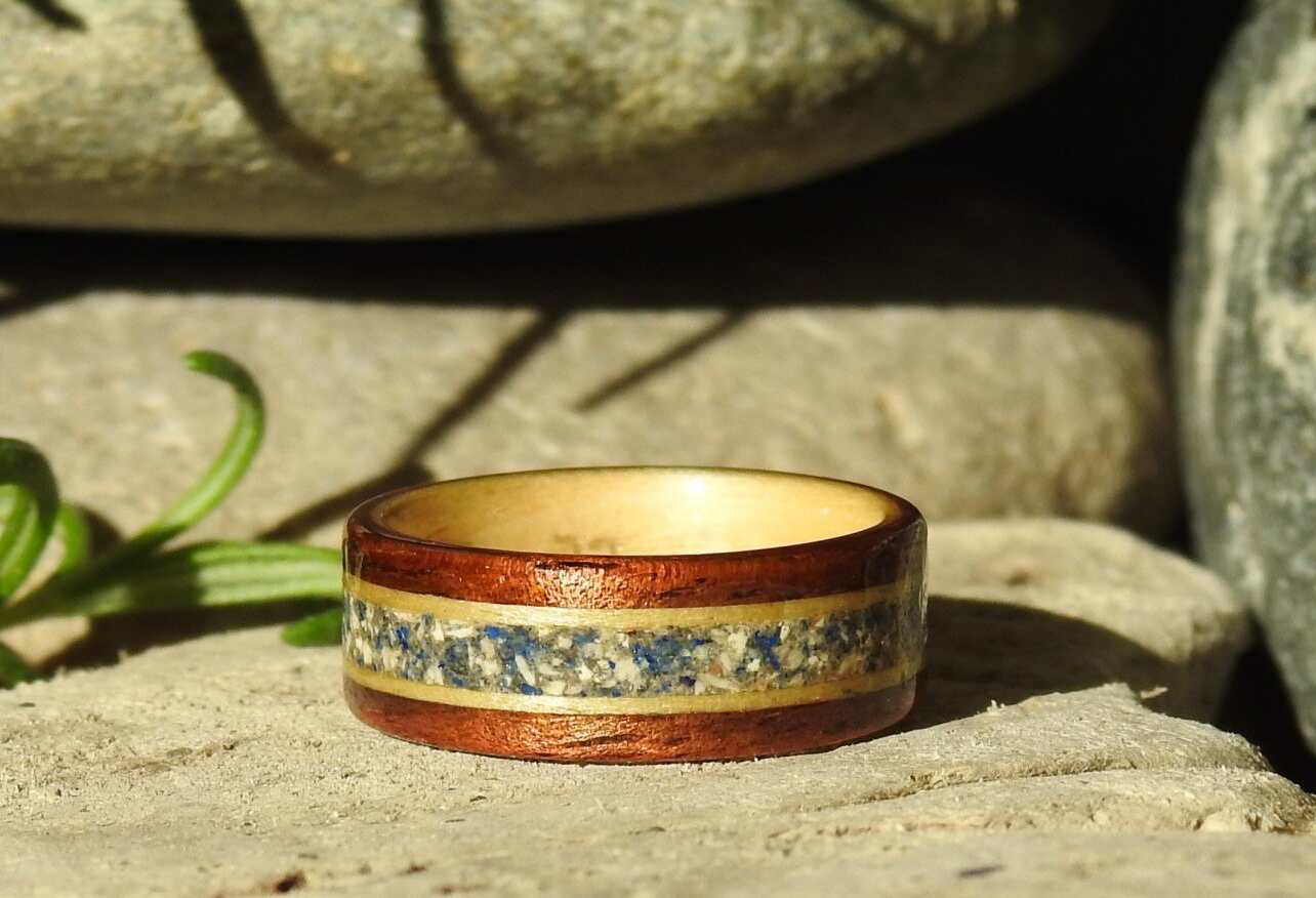 Memorial ring made of wood with inlaid ashes, resting on driftwood with a sprig of rosemary in the background.
