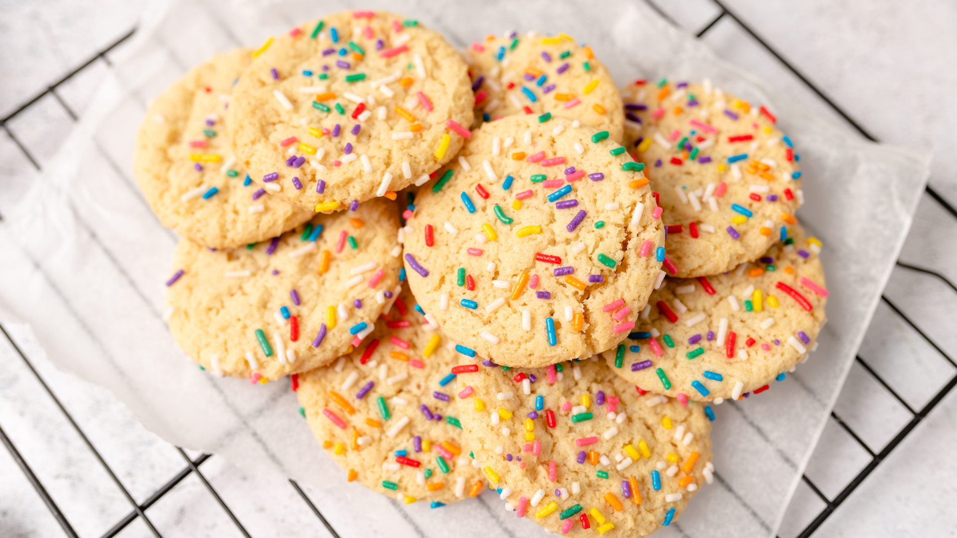 A small pile of round, golden-brown sugar cookies with colorful sprinkles resting on parchment paper atop a wire rack.
