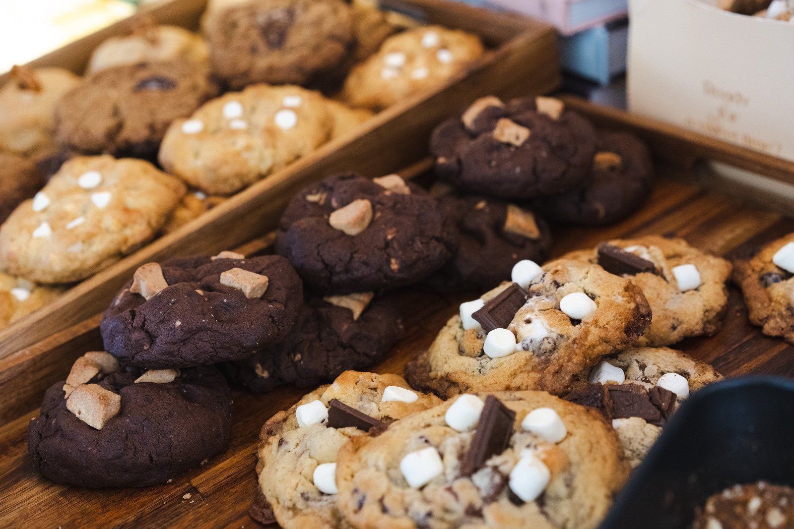 Rows of freshly baked chocolate chip and s'mores cookies arranged in wooden display trays.