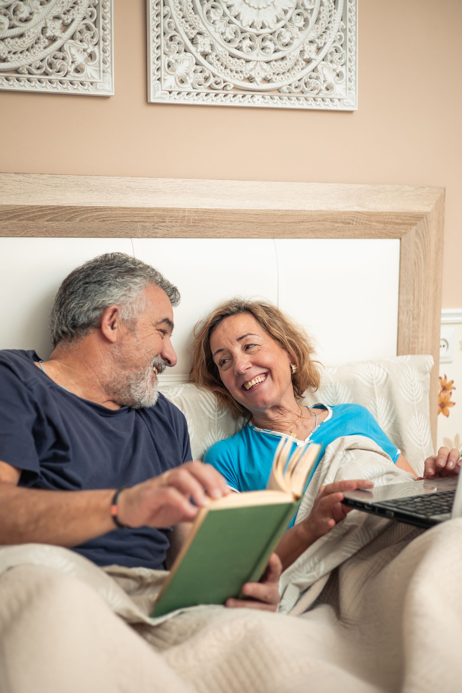 Couple smiles while reading a book and using a laptop in bed.
