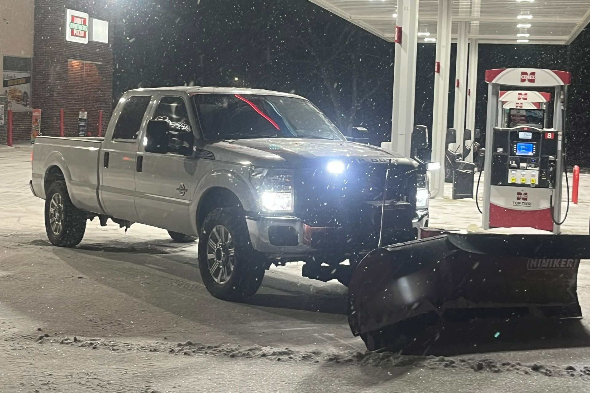 Pickup truck at a gas station at night, headlights on, beside a snowplow blade and fuel pump