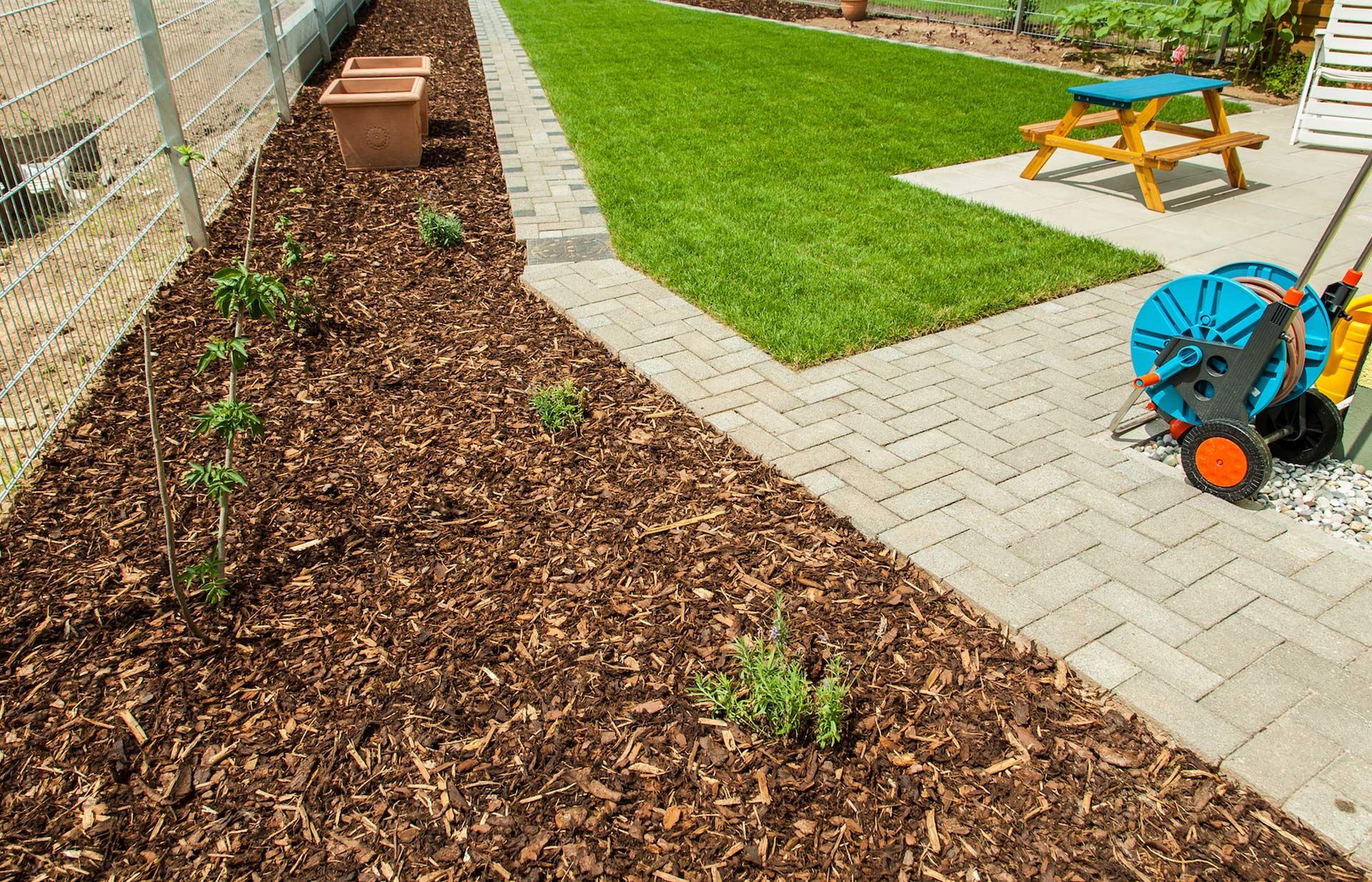 Mulched garden bed beside a paved patio with a blue hose reel and wooden bench in a backyard