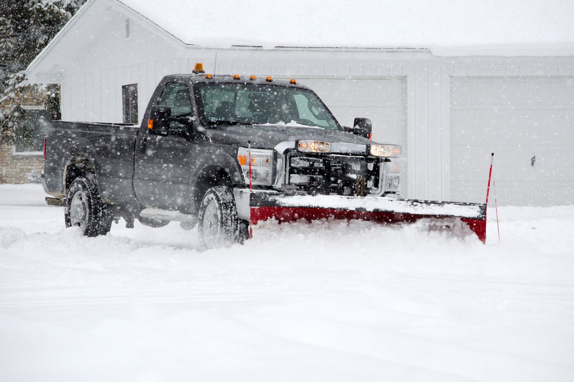 Black pickup with snow plow driving through deep snow in front of a house