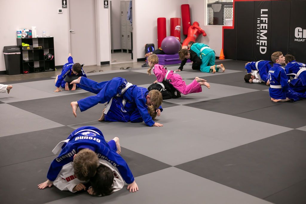 Children in blue martial arts uniforms practicing drills on mats in a gym, with an instructor in the background